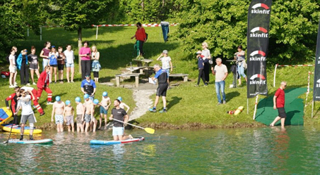 Eine Gruppe von Menschen steht am See, einige in Badekleidung und Helmen. Eine Person paddelt auf einem blauen Brett. In der Nähe befinden sich Werbebanner.