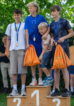 Fünf Jungen stehen auf einem Podest. Drei haben Medaillen um den Hals. Ein Junge hat orangefarbene Taschen an den Schultern. Die Zahlen 1, 2 und 3 stehen auf dem Podest.