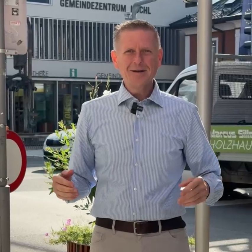 Ein Mann in einem blau gestreiften Hemd und khakifarbener Hose steht auf einer Stadtstraße, lächelt und gestikuliert, während er spricht. Hinter ihm befindet sich ein Gebäude mit dem Schild 'Gemeindezentrum' und ein geparkter Lieferwagen.