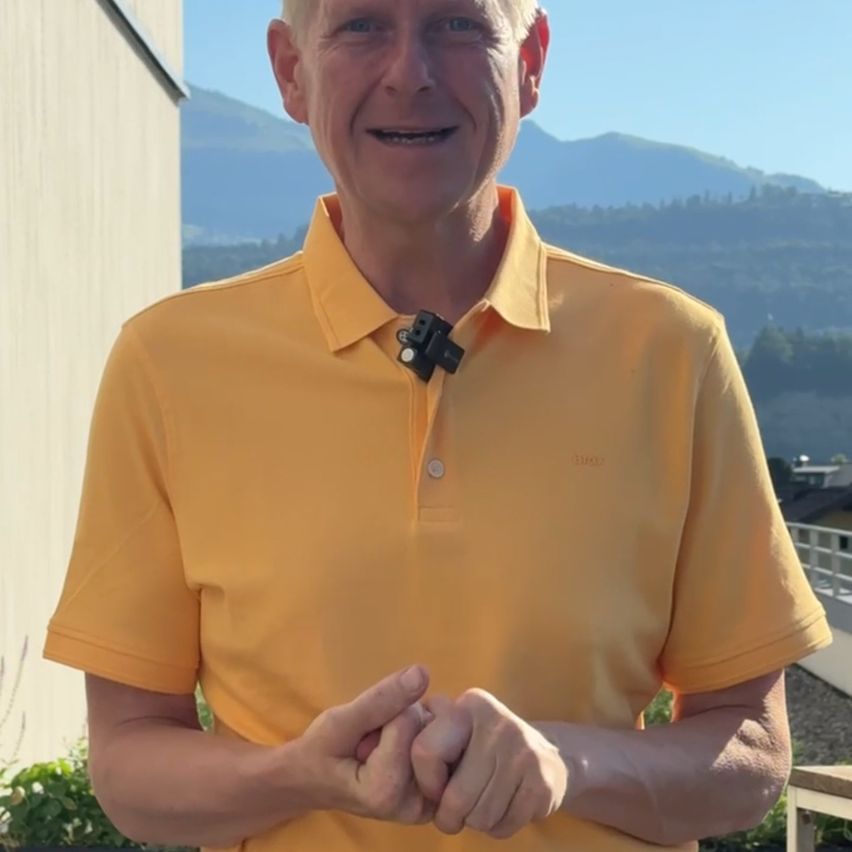 Ein Mann in einem gelben Polohemd mit einer schwarzen Kamera am Kragen steht draußen, lächelt und posiert für ein Foto mit Bergen im Hintergrund.