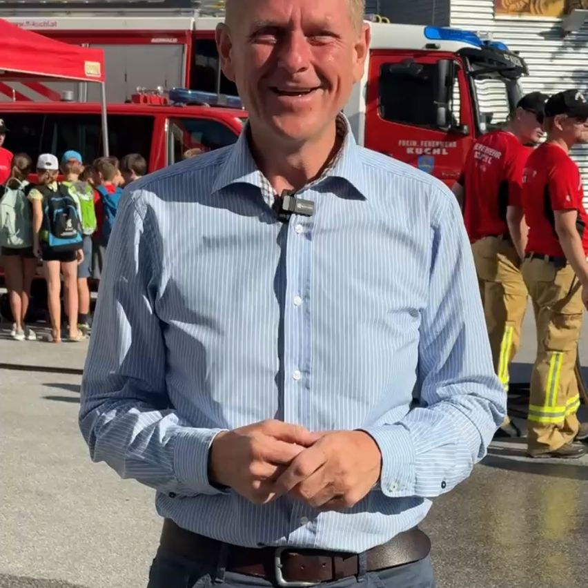 Ein Mann in einem hellblauen Hemd steht lächelnd mit gefalteten Händen vor ihm, in der Nähe eines Feuerwehrautos und einer Gruppe von Feuerwehrleuten.