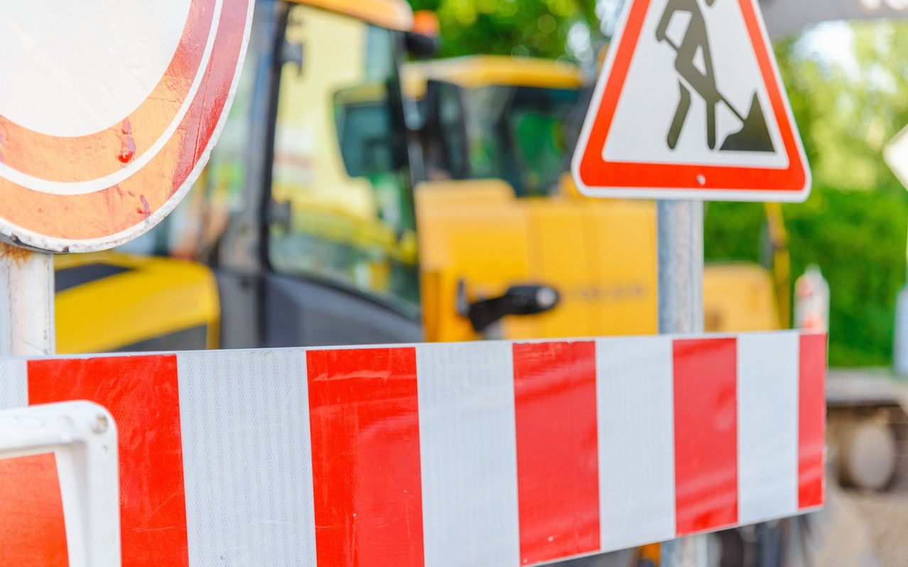 Ein Straßenbaufahrzeug ist geparkt, mit einem rot-weiß gestreiften Barriereband und einem dreieckigen Warnzeichen mit einer Figur, die eine Schaufel hält.