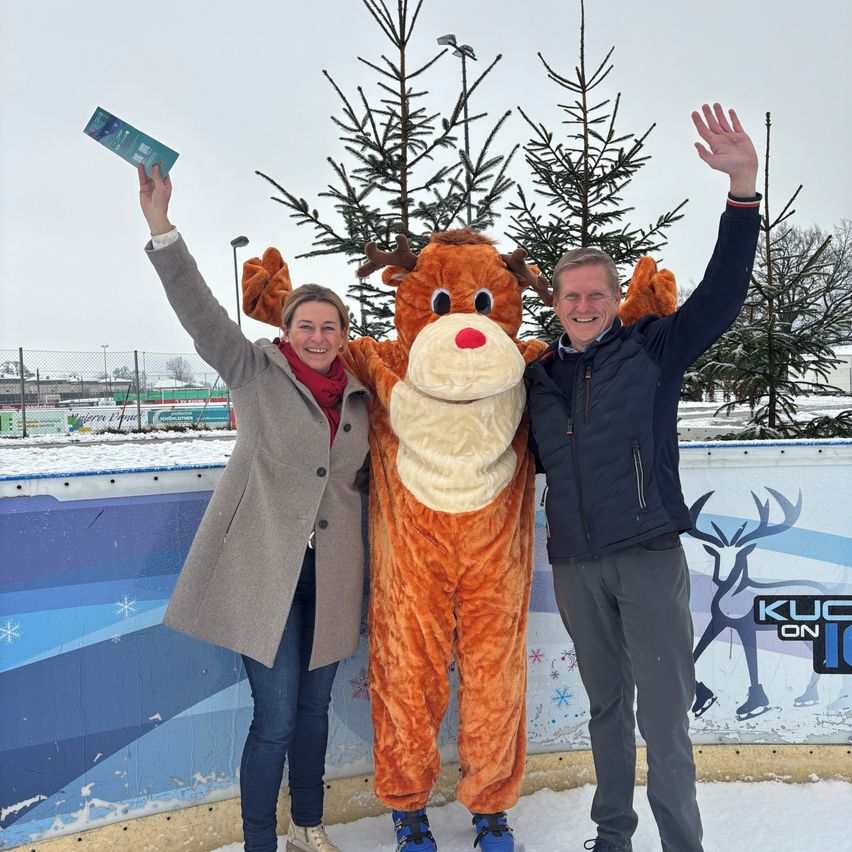 Eine Frau, ein Mann und ein Maskottchen mit roter Nase posieren vor einer Eisbahn mit schneeiger Kulisse.