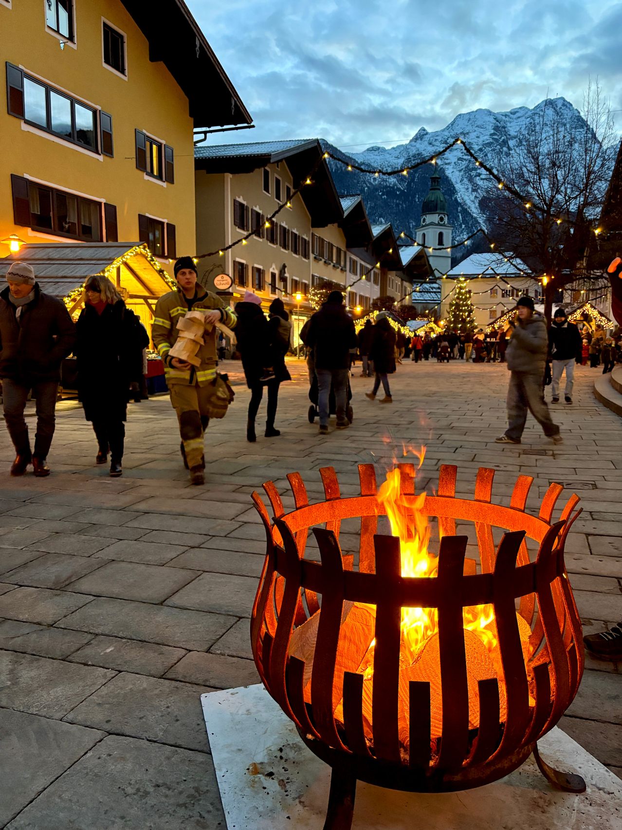Eine Feuerstelle, umgeben von einem Metallgitter, steht in der Mitte eines Stadtplatzes, wo viele Menschen herumlaufen. Im Hintergrund sind schneebedeckte Berge zu sehen.