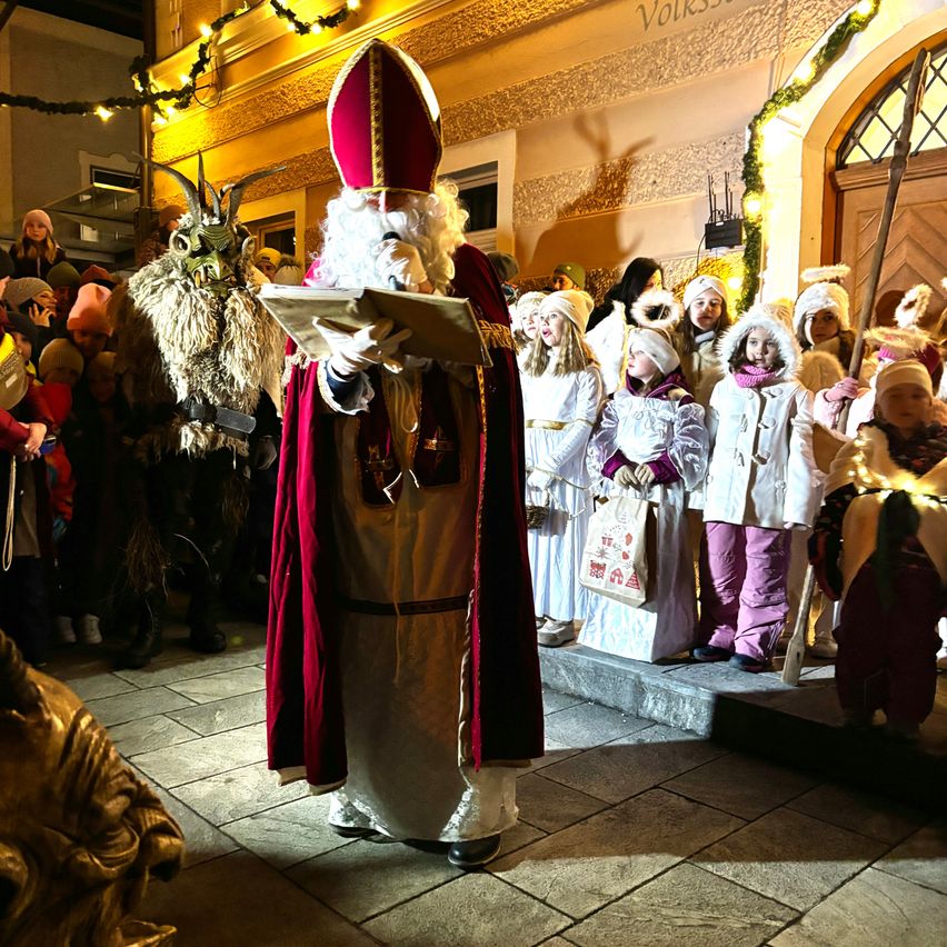 Ein Mann, der als St. Nikolaus verkleidet ist, liest ein Buch vor einer Menschenmenge von Kindern, einige tragen Masken und haben Taschen bei sich.