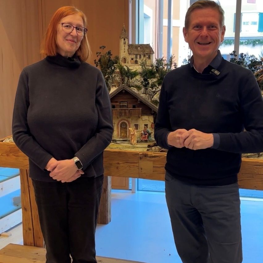 Ein Mann und eine Frau stehen lächelnd vor einem Holztisch und posieren für ein Foto. Hinter ihnen befindet sich eine Miniaturszene eines Hauses mit einer Kirche.
