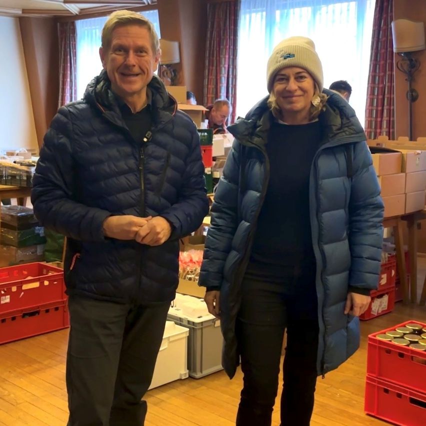 Ein Mann und eine Frau stehen nebeneinander in einem Raum mit Holzboden. Sie lächeln beide. Hinter ihnen befinden sich Lagerboxen und andere Gegenstände. An der Wand steht eine Lampe.