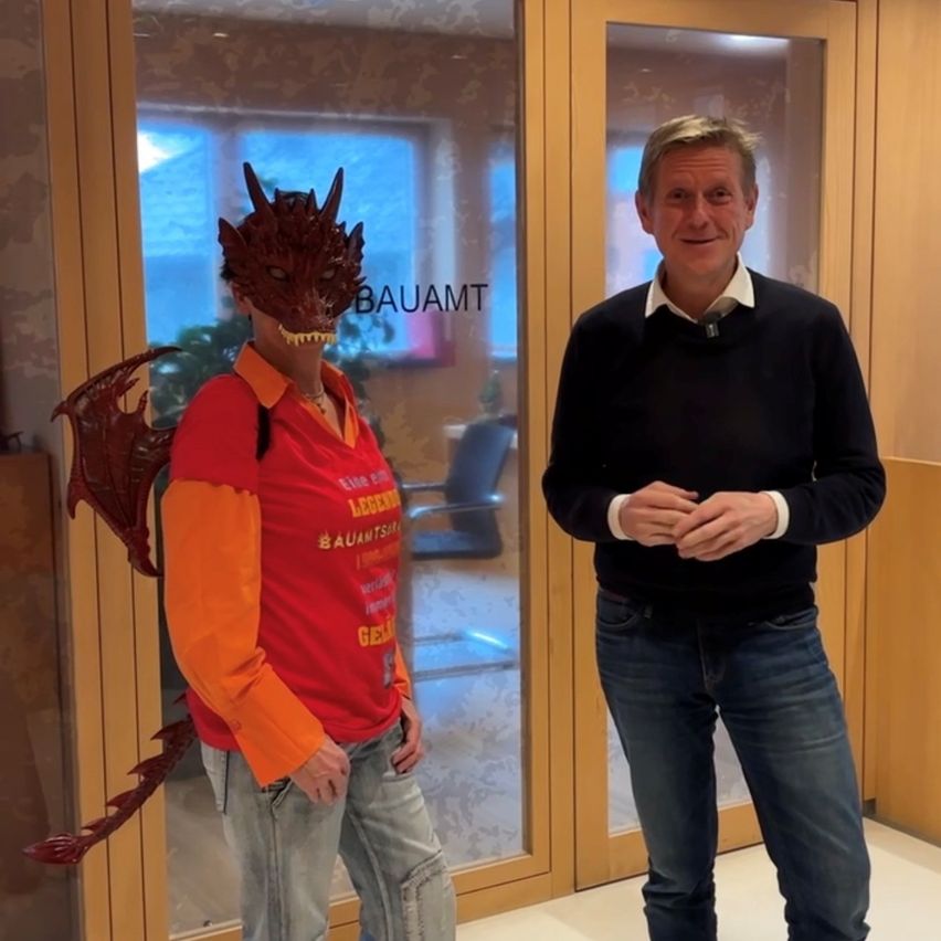 Zwei Männer stehen vor einer Glastür. Einer trägt eine Drachenmaske und ein rotes Shirt, während der andere einen schwarzen Pullover und Jeans trägt. Dahinter befindet sich ein Raum mit einem Schreibtisch und einer Pflanze.