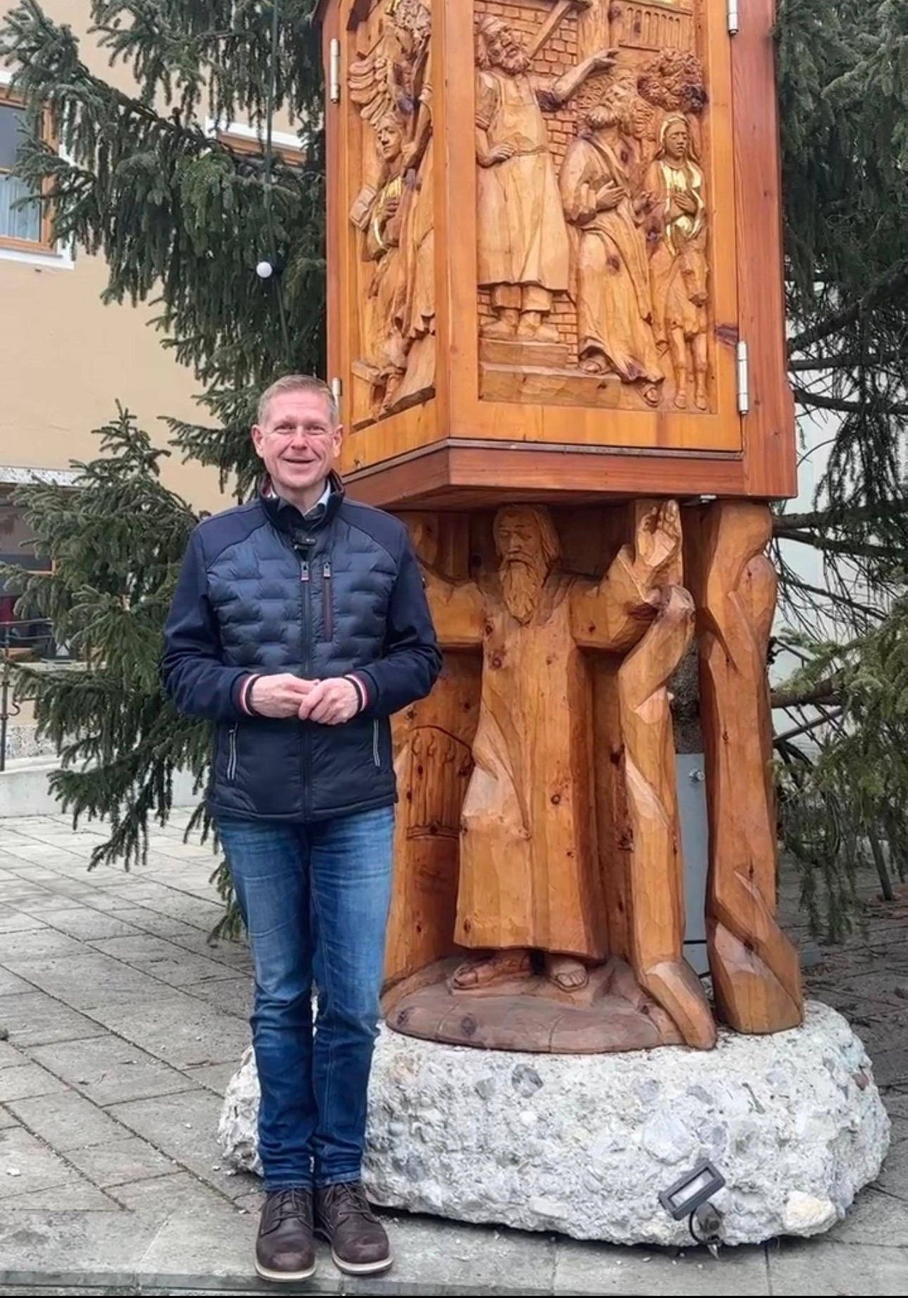 Ein Mann steht neben einer großen hölzernen Skulptur einer Figur mit einem Stab, die auf einem Steinsockel mit Schnitzereien darüber steht. Hinter ihm sind ein Baum und ein Gebäude zu sehen.