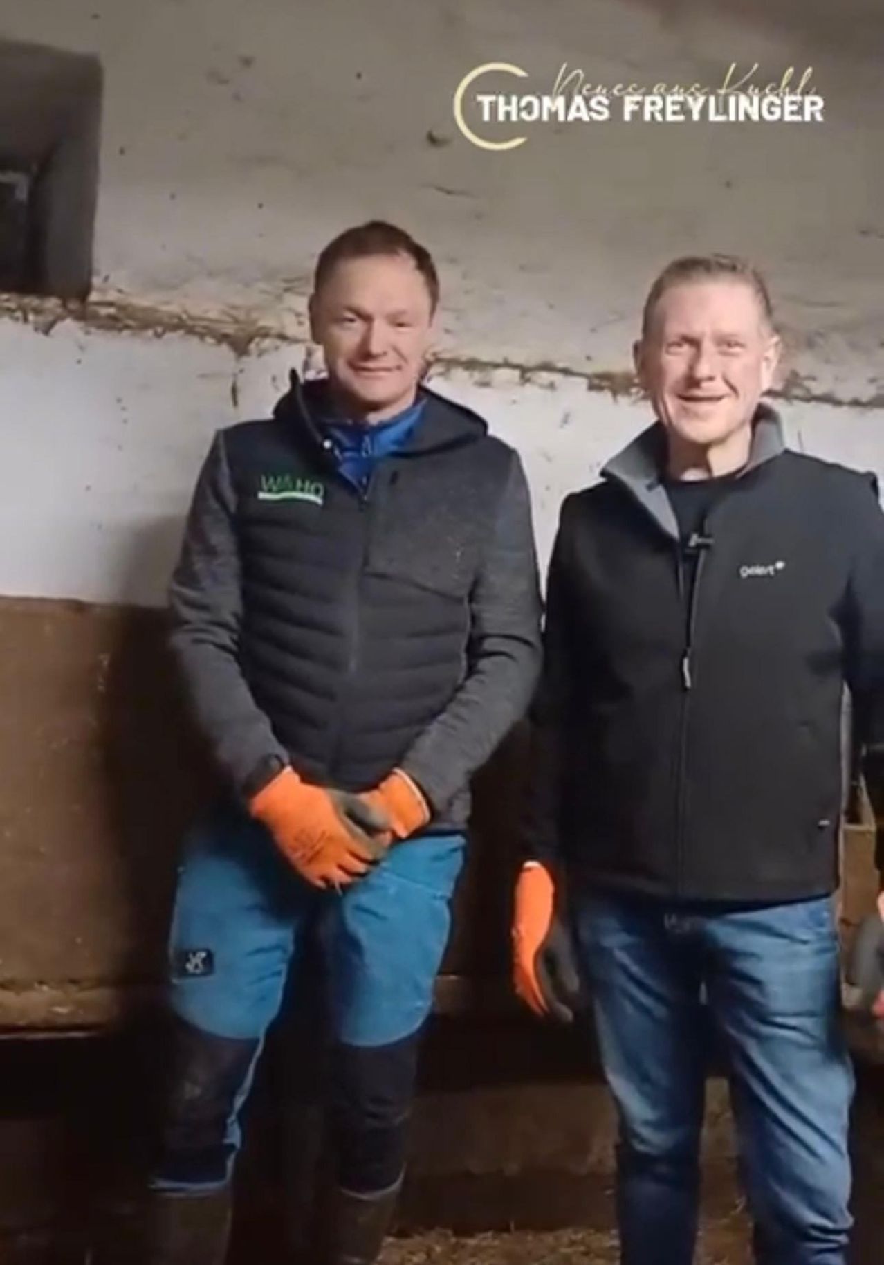 Zwei Männer in Arbeitskleidung, lächelnd vor einer weißen Wand stehend. Beide tragen orange Handschuhe und Jacken mit Logos auf der linken Brust.