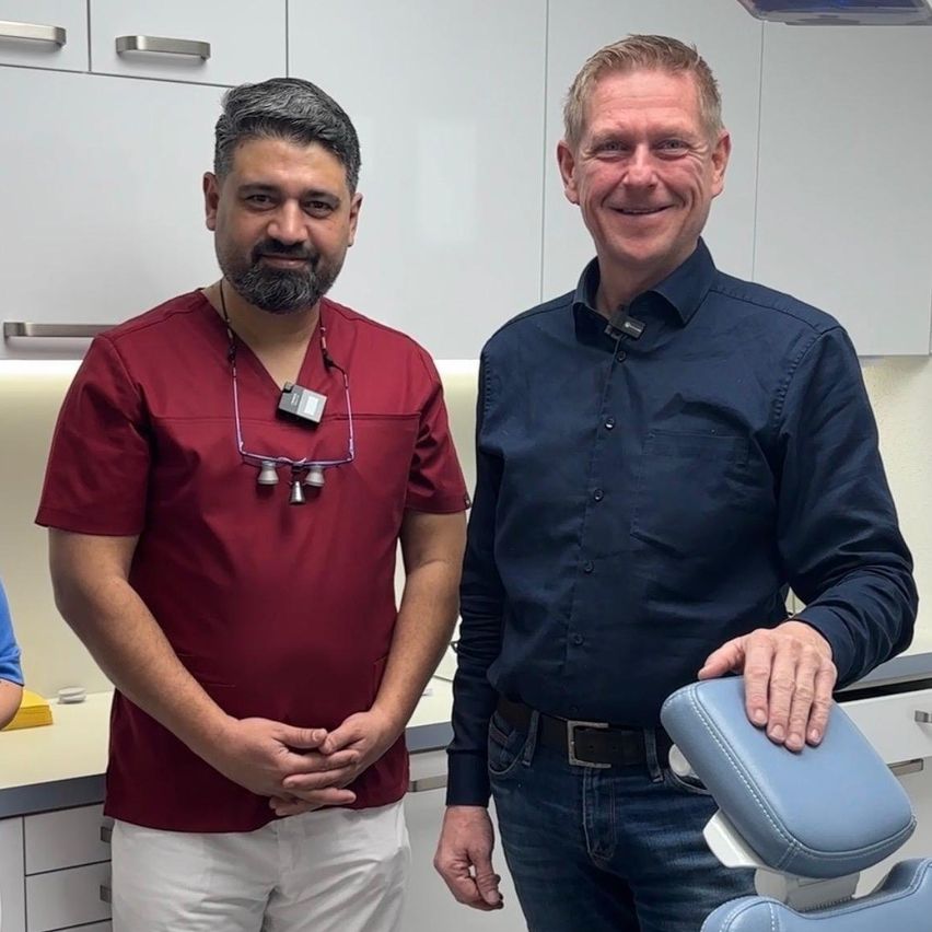 Zwei Männer stehen in einer Zahnarztpraxis. Einer trägt ein rotes Scrub, der andere ein blaues Hemd. Beide lächeln in die Kamera. Hinter ihnen befinden sich weiße Schränke.