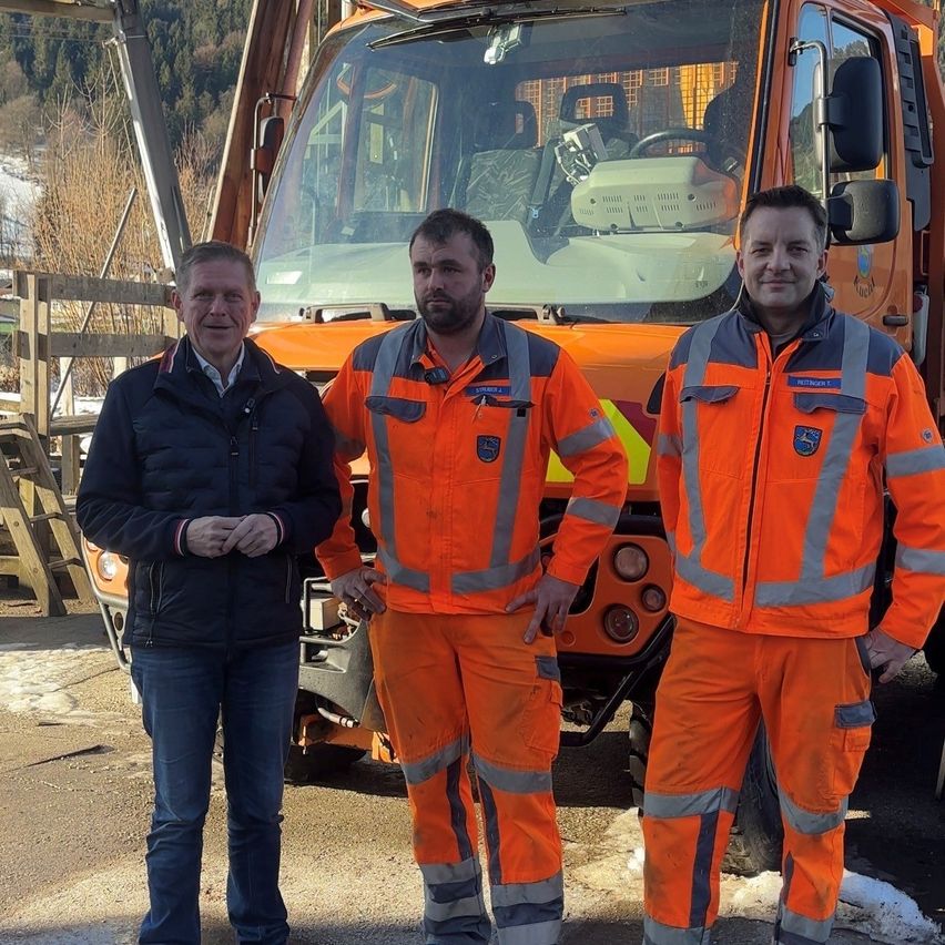 Drei Männer in Sicherheitsanzügen stehen vor einem orangefarbenen Fahrzeug mit verschneiten Bergen im Hintergrund.