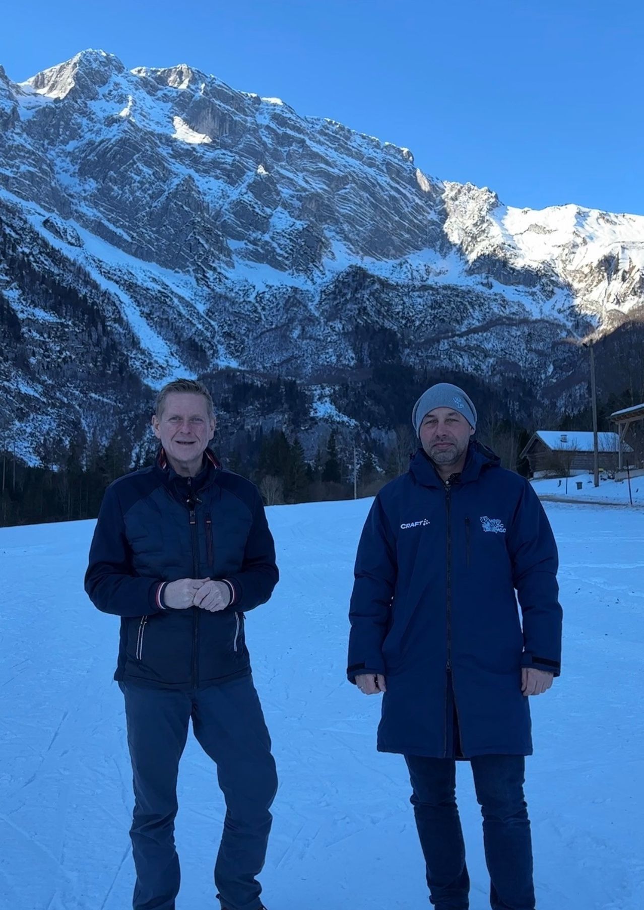 Zwei Männer stehen auf einem verschneiten Berg und lächeln. Einer trägt einen blauen Mantel mit Craft- und Snowflake-Logos. Hinter ihnen verschneite Berge und ein Holzhaus.