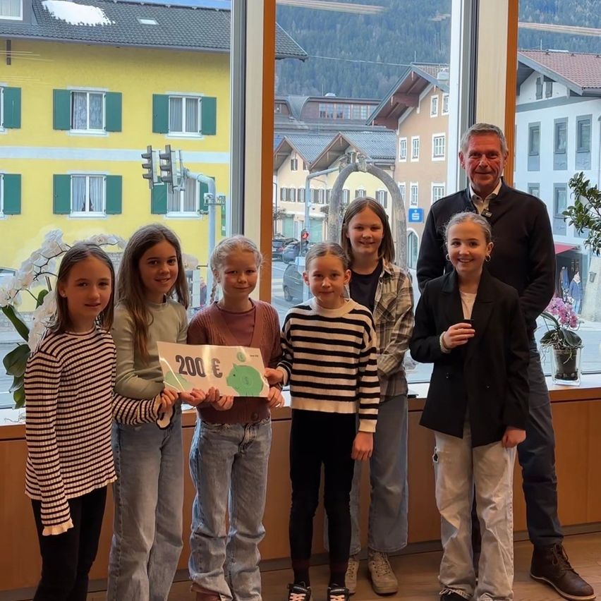 Fünf Mädchen und zwei Männer stehen lächelnd zusammen drinnen und halten ein Schild mit 200€. Dahinter zeigt ein Fenster ein Bergdorf mit Gebäuden und Ampeln.