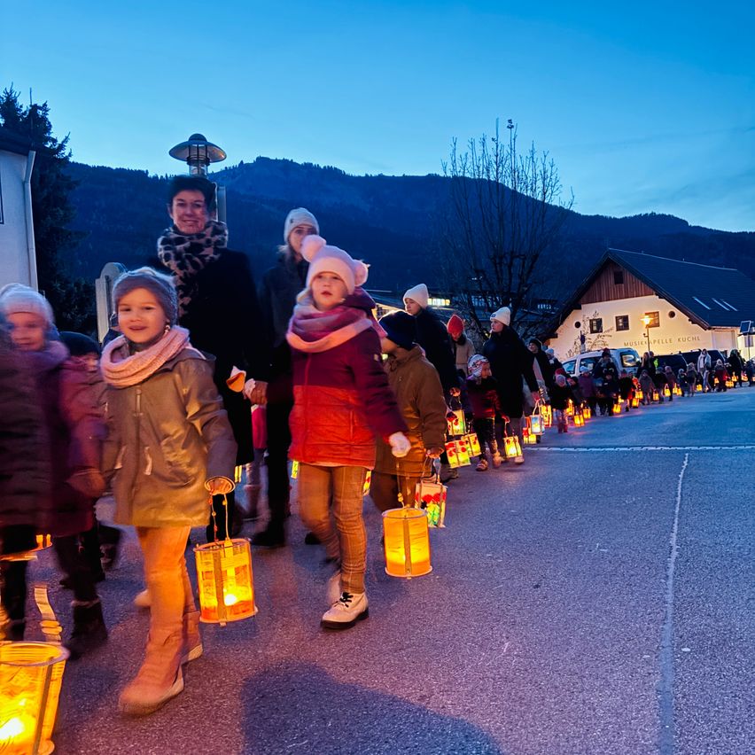 Kinder in Winterkleidung gehen bei Dämmerung eine Straße entlang und halten Laternen. Dahinter laufen Erwachsene, und Berge sind im Hintergrund.