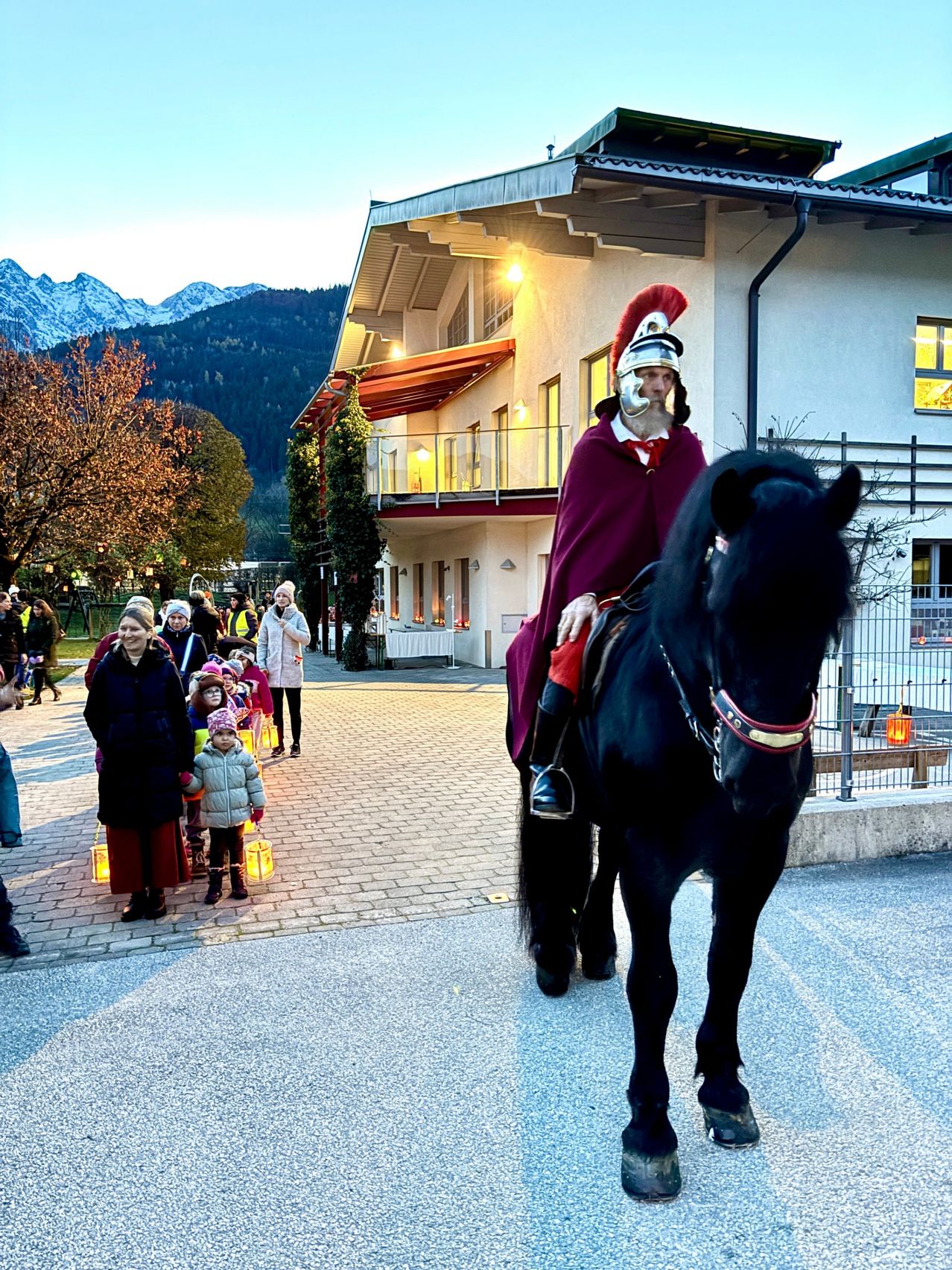 Ein Mann in römischer Soldatenkleidung reitet ein schwarzes Pferd vor einem Gebäude mit verschneiten Bergen im Hintergrund.