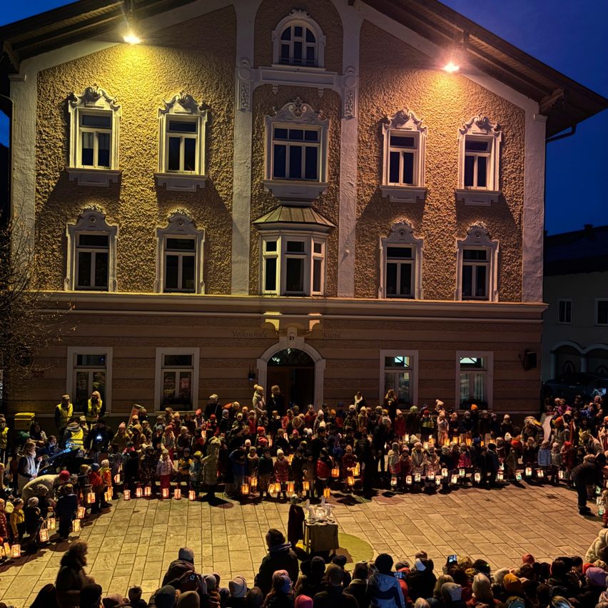 Eine große Menschenmenge versammelt sich mit Laternen in der Hand vor einem großen Gebäude mit vielen Fenstern.