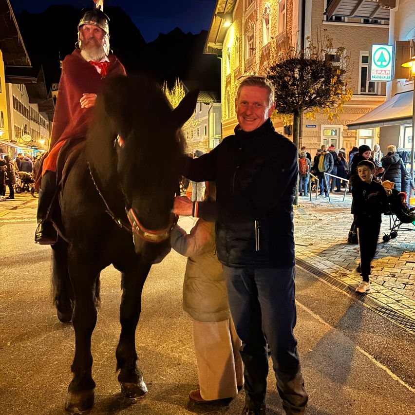 Ein Mann in mittelalterlicher Kleidung reitet ein schwarzes Pferd, während ein anderer Mann neben ihm lächelt. Sie befinden sich auf einer Kopfsteinpflasterstraße mit Gebäuden und Menschen im Hintergrund.
