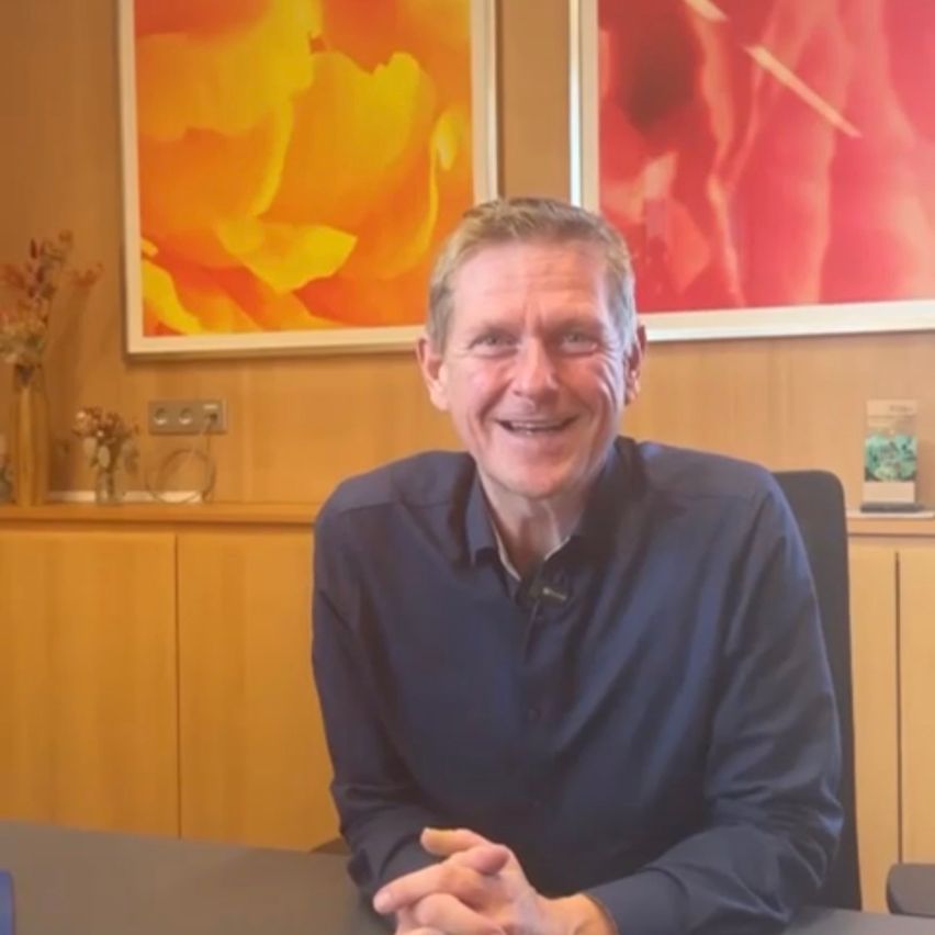 Ein älterer Mann mit einem Lächeln sitzt in einem Bürostuhl vor einem Schreibtisch, mit zwei abstrakten Gemälden an der Wand hinter ihm.