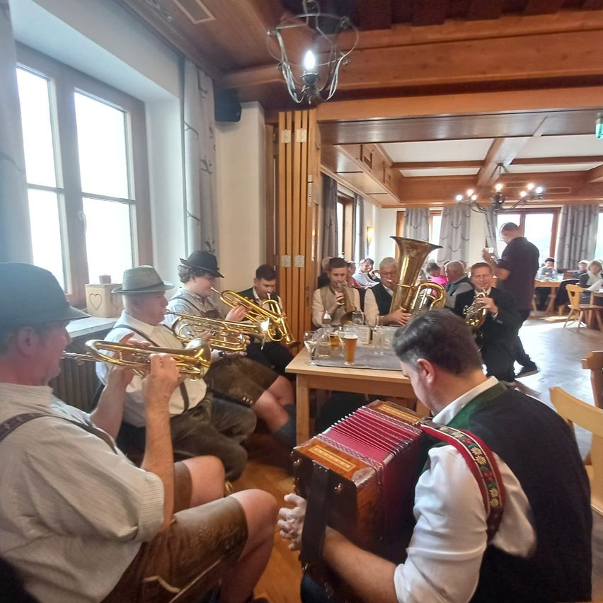 Eine Gruppe von Musikern spielt in einem Restaurant Instrumente, darunter Tuba und Akkordeon. Sie sitzen um einen Tisch mit Gläsern Bier herum. An der Wand ist ein Lautsprecher angebracht.