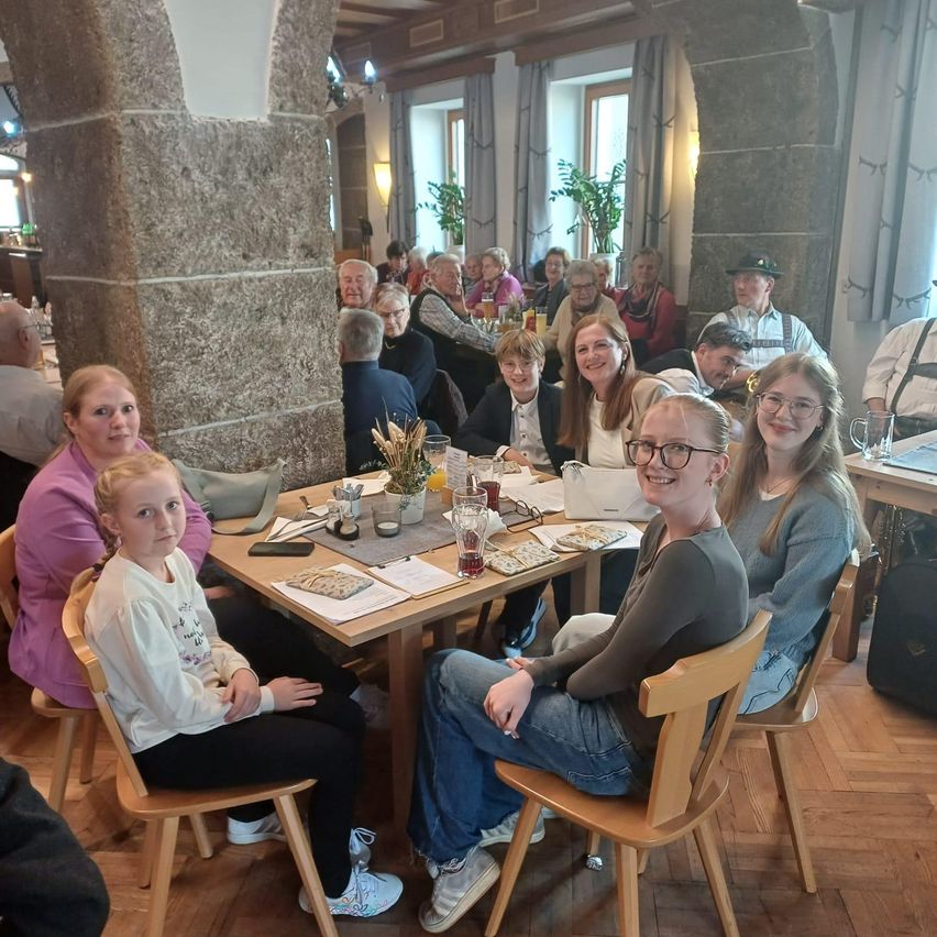 Eine Gruppe von Menschen, darunter Erwachsene und Kinder, sitzt um einen Tisch in einem Restaurant. Auf dem Tisch befinden sich verschiedene Gegenstände wie Gläser, eine Handtasche und ein Notizbuch. Der Raum hat Steinsäulen und Vorhänge.