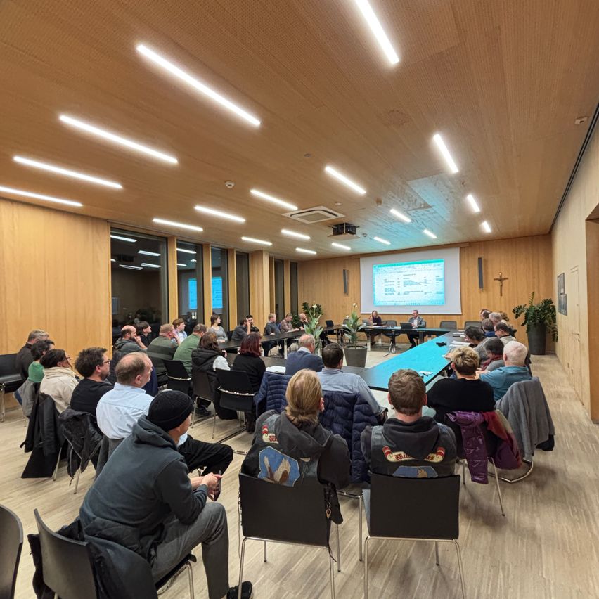 Eine Gruppe von Menschen sitzt in einem Raum mit Holzwänden und hört einer Präsentation zu, die auf einer Projektionsleinwand angezeigt wird. Der Raum hat einen Holzboden und wird durch Einbauleuchten beleuchtet.