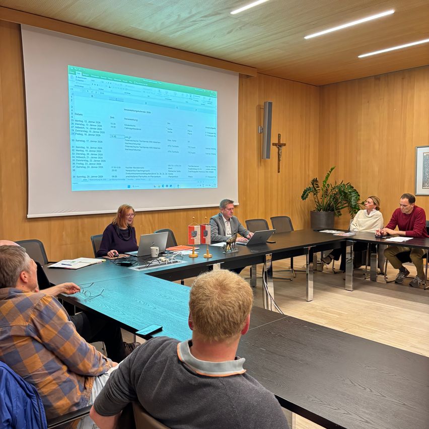 Eine Besprechung mit einer Gruppe von Personen, die um einen Tisch in einem Konferenzraum herum sitzen. Eine Präsentation wird auf einer Projektionsleinwand gezeigt.