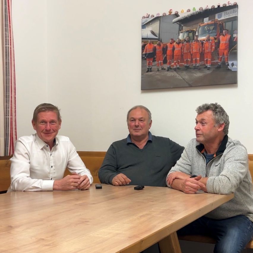 Drei Männer sitzen an einem Holztisch, mit einem Foto von Arbeitern in orangefarbenen Uniformen hinter ihnen.