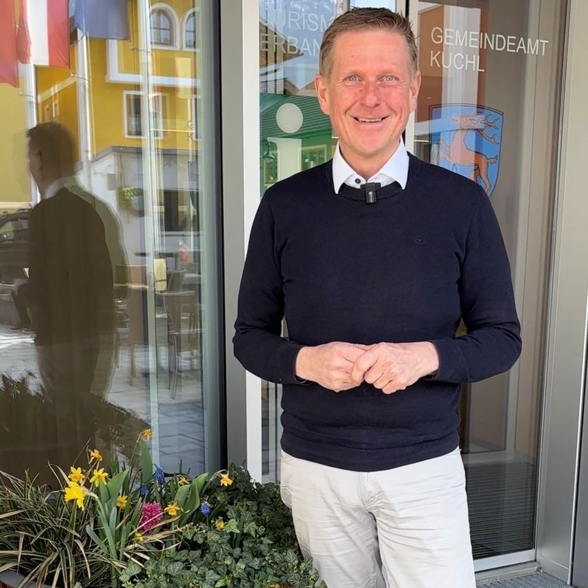 A man in a navy sweater and khaki pants stands outside a building, smiling. Behind him is a glass door with the sign Gemeindeamt Kuchl. A man in a suit is reflected in the glass.