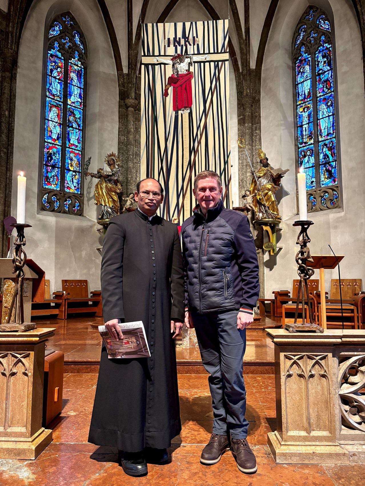 Two men stand in a church with candles and statues behind them. The man on the left wears a black robe and holds a book. The man on the right wears a blue jacket and smiles.
