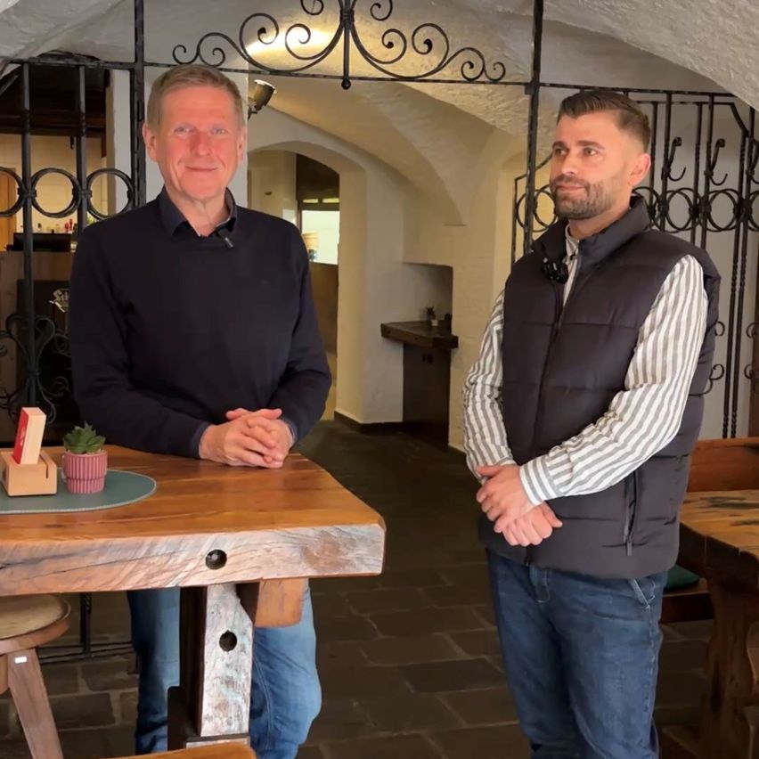Two men stand in a room with a wooden table and a plant. One man is smiling and has his hands clasped together, while the other is standing with his hands on his hips. Behind them is a black metal gate.