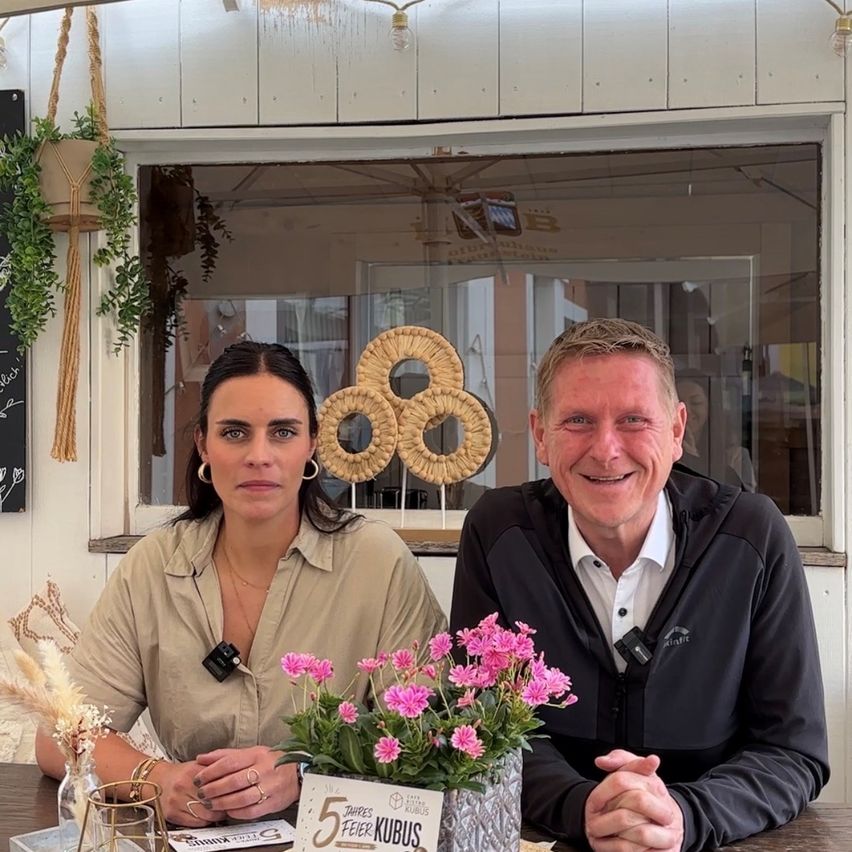 Ein Mann und eine Frau sitzen an einem Tisch mit Blumen. Der Mann lächelt. Hinter ihnen ist ein Fenster mit Blick auf das Freie.