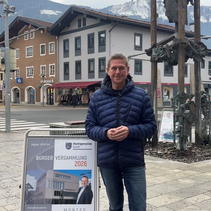 Ein Mann in einer blauen Jacke steht vor einem Schild, das eine Burgerversammlung 2026 ankündigt. Dahinter befinden sich Gebäude, Berge und eine Statue.
