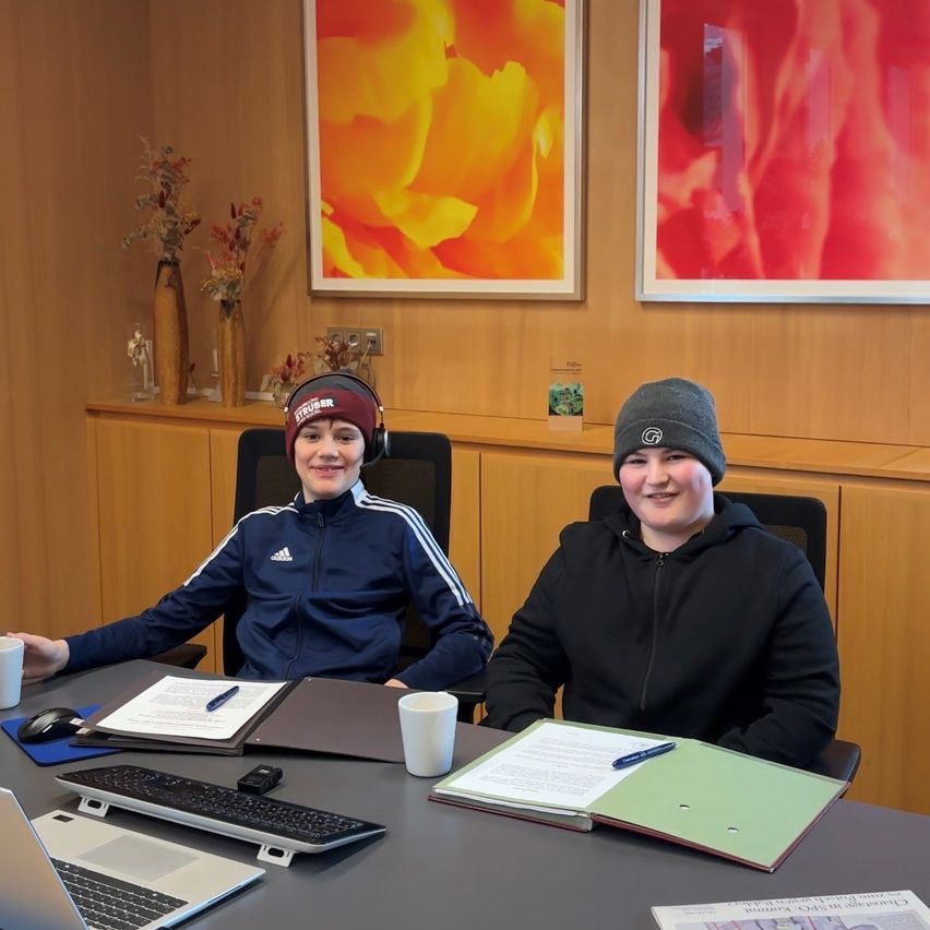 Zwei Personen sitzen in einem Büro an einem Schreibtisch und tragen Wintermützen. Einer hält eine Tasse, der andere einen Stift. Hinter ihnen hängen zwei abstrakte Gemälde und Holzschränke.