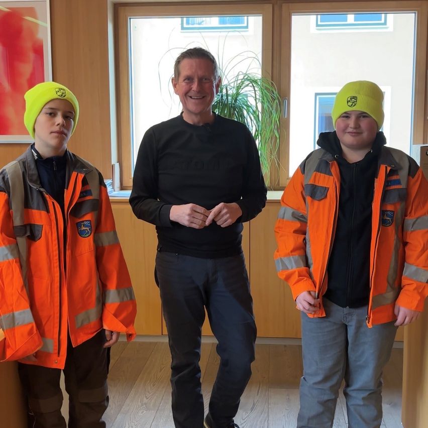 Drei Personen stehen in einem Raum, zwei tragen orangefarbene Jacken und ein Mann in einem schwarzen Hemd lächelt. Hinter ihnen zeigt ein großes Fenster eine Topfpflanze.