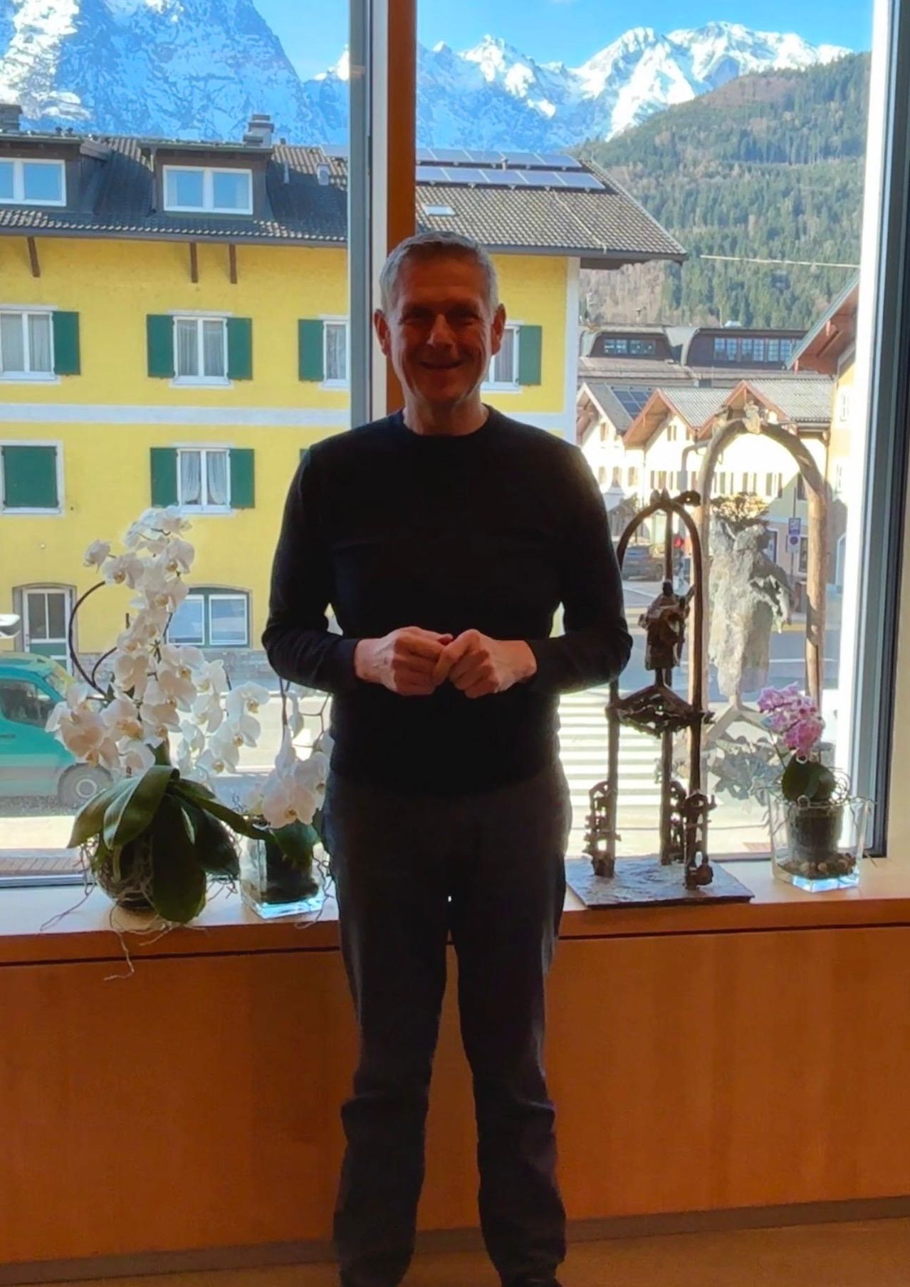 Ein Mann steht in einem Raum mit einer gelben Wand, lächelt und posiert für ein Foto. Vor ihm stehen zwei Vasen mit weißen Blumen. Hinter ihm befindet sich ein Fenster mit Blick auf ein Gebäude und ein Auto.