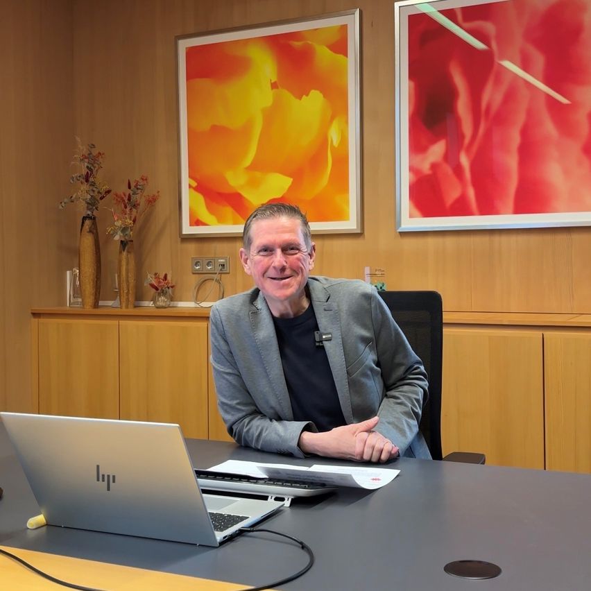 Ein Mann sitzt mit einem Laptop vor sich lächelnd an einem Schreibtisch. Hinter ihm hängen zwei große Gemälde an der Wand.