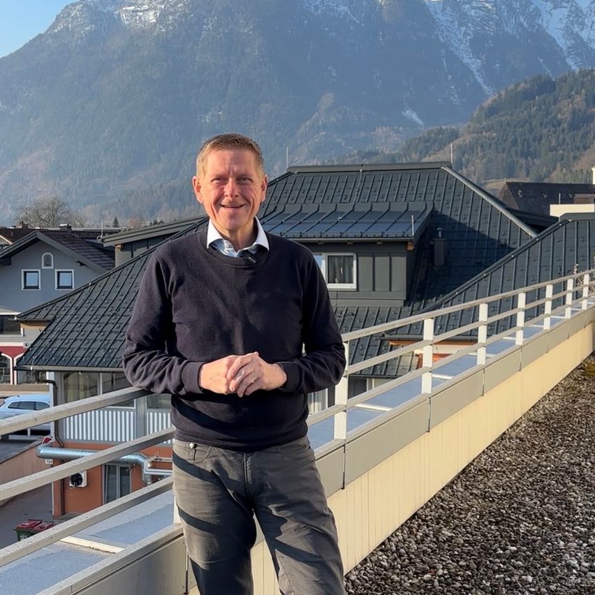A man in a black sweater and jeans poses for a photo on a balcony with a mountain view.