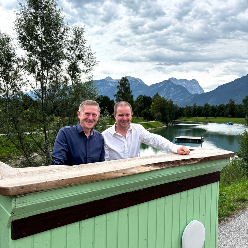 Zwei Männer stehen lächelnd hinter einer grünen Bartheke, mit einem See und Bergen im Hintergrund.