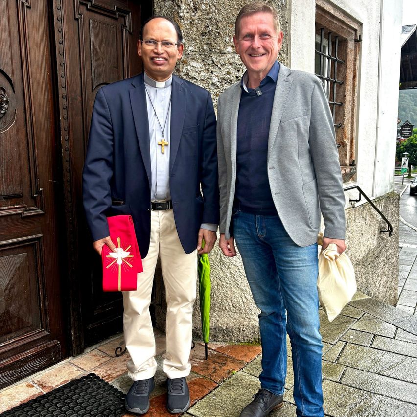 Zwei Männer stehen vor einer Kirche, einer in geistlicher Kleidung mit einer roten Stola mit einem Kreuz, der andere in lässiger Kleidung mit einem grünen Regenschirm.