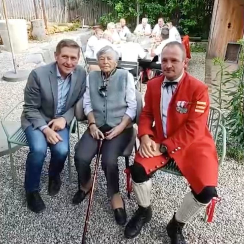 Drei Männer posieren für ein Foto in einem Garten. Ein älterer Mann hält einen Stock, und zwei Männer in formeller Kleidung sitzen neben ihm. Sie sind von einer kleinen Menge umgeben.