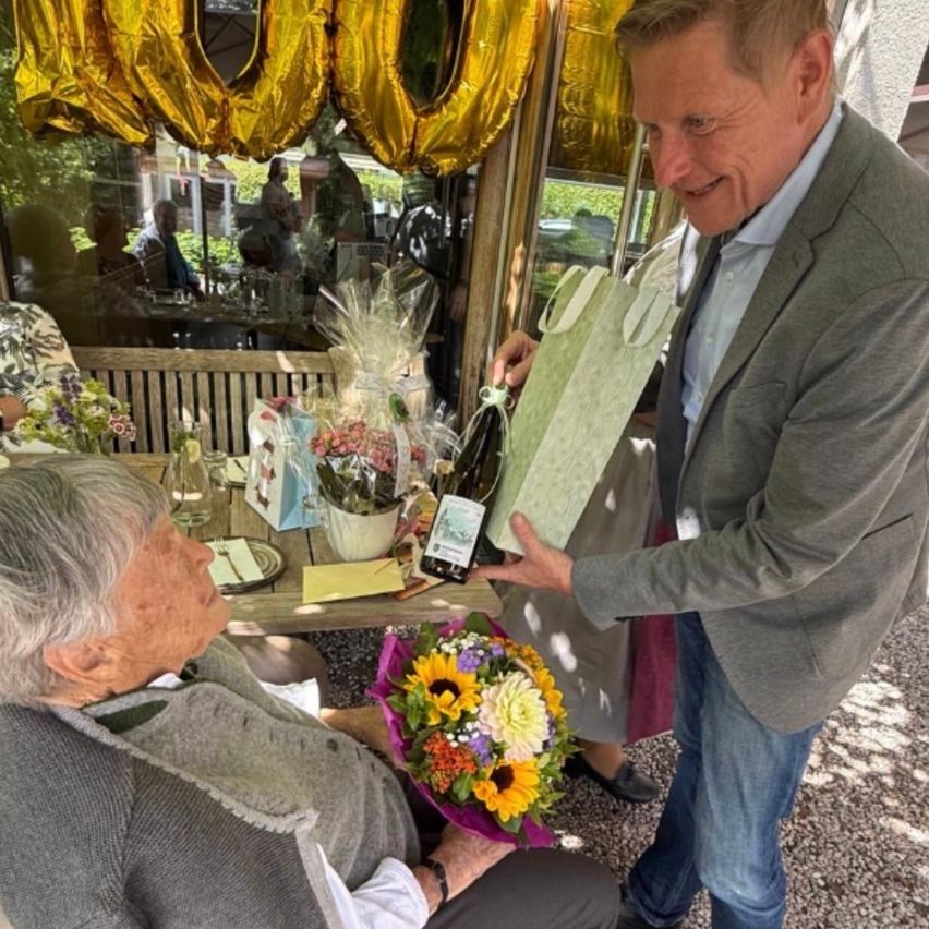 Ein älterer Mann überreicht einer älteren Frau in einem Garten einen Blumenstrauß und eine Flasche Champagner.
