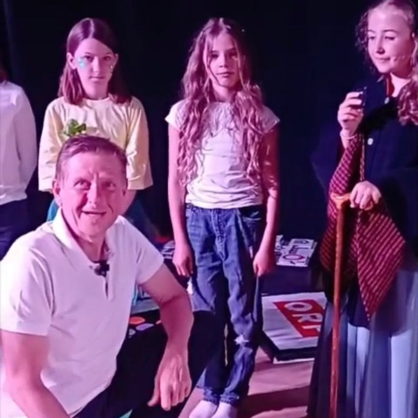 A group of children and an adult man are on stage. They appear to be performing a play. The man is smiling and sitting.