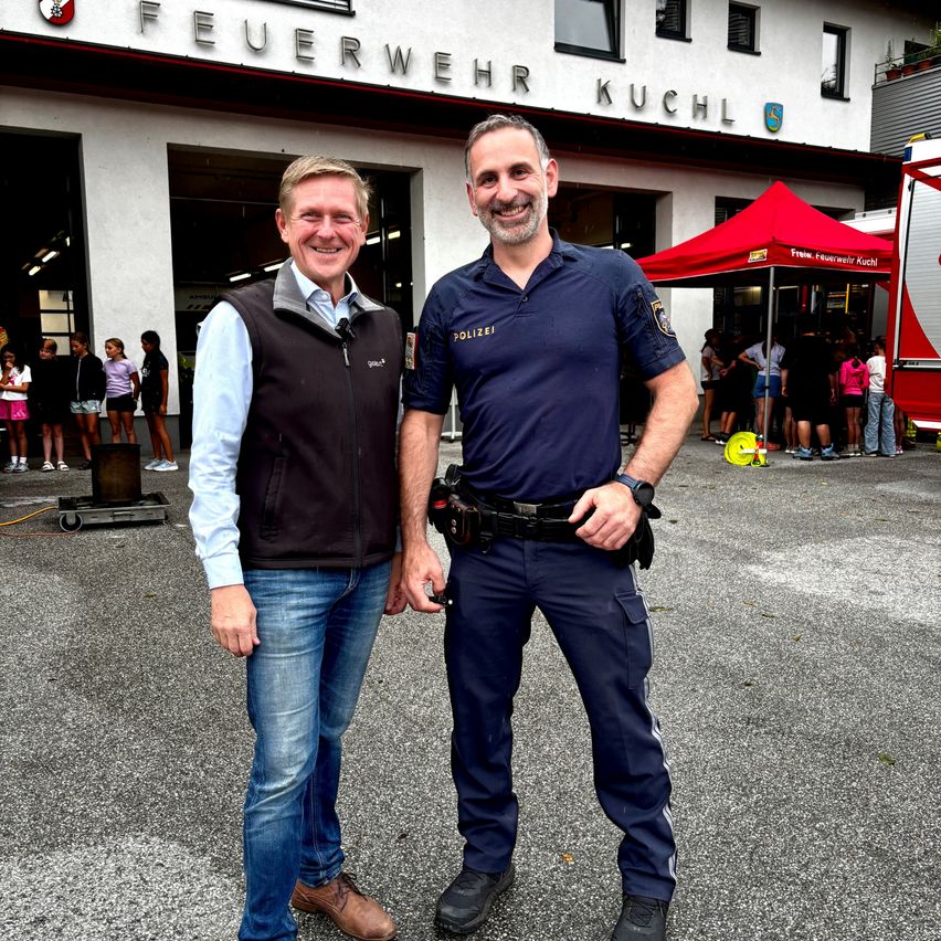 Two men stand in front of a fire station, one in a vest and the other in a blue uniform. Behind them, people gather near a red tent and a fire truck.