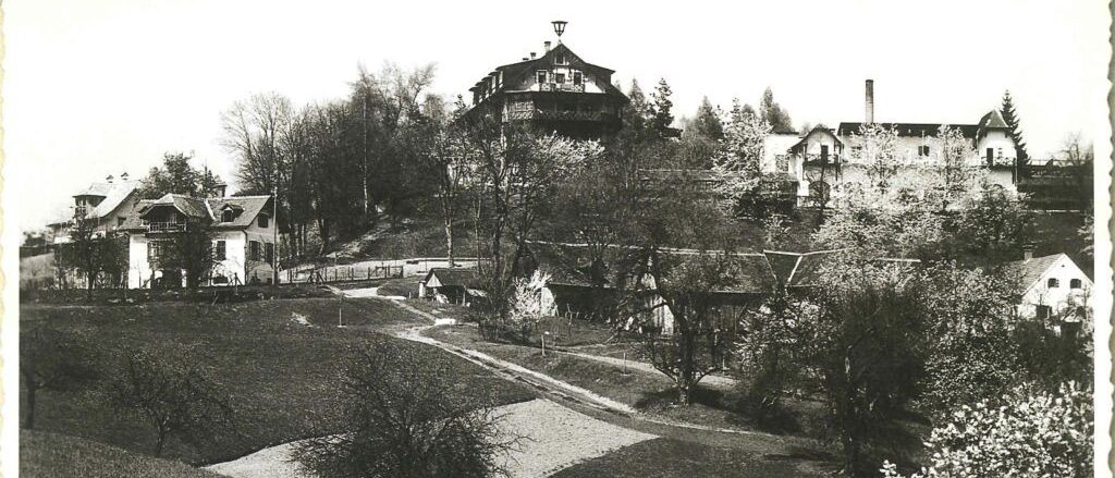 Ein Schwarz-Weiß-Bild zeigt einen Hügel mit einem großen Gebäude, möglicherweise einem Sanatorium, umgeben von Bäumen und einem gewundenen Pfad. Kleinere Häuser sind ebenfalls auf dem Hügel sichtbar.