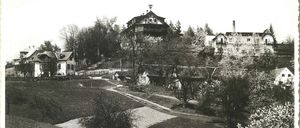 Ein Schwarz-Weiß-Bild zeigt einen Hügel mit einem großen Gebäude, möglicherweise einem Sanatorium, umgeben von Bäumen und einem gewundenen Pfad. Kleinere Häuser sind ebenfalls auf dem Hügel sichtbar.