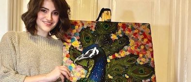 Eine Frau steht neben einer Tafel mit einem Pfauengemälde und lächelt.