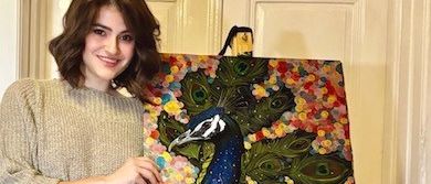 Eine Frau in einem beigen Kleid und schwarzen Strumpfhosen steht hinter einem Staffelei mit einem farbenfrohen Gemälde eines Pfaus.