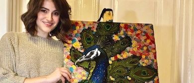 Eine Frau steht neben einem farbenfrohen Gemälde eines Pfaus, lächelt und hält den Staffelei.