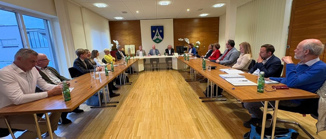 Eine Besprechung im Gange mit Teilnehmern, die um einen Konferenztisch in einem Raum mit Holzboden und einer mit einem Wappen geschmückten Wand sitzen.